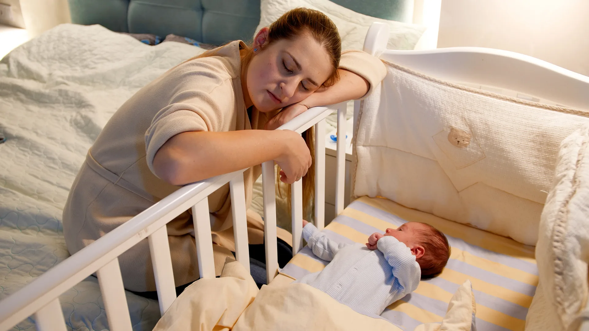 Mama s hlavou opretou o hranu kolísky, v ktorej leží bdiace dieťa.
